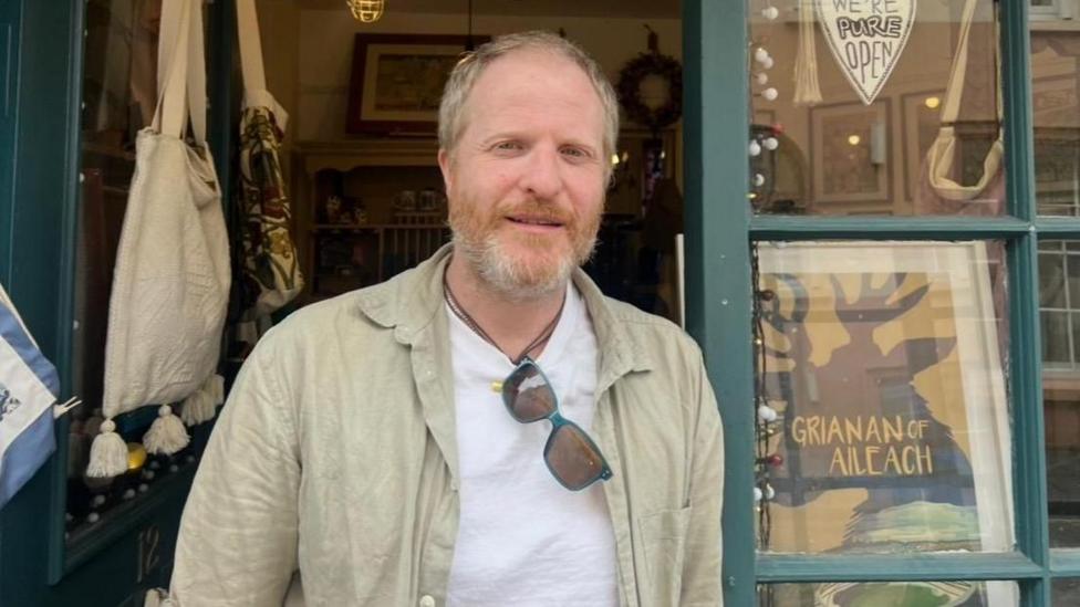 Pauric Lawne is stadning in a shop doorway, wearing a white tshirt and beige coat. He has short blond hair and beard.