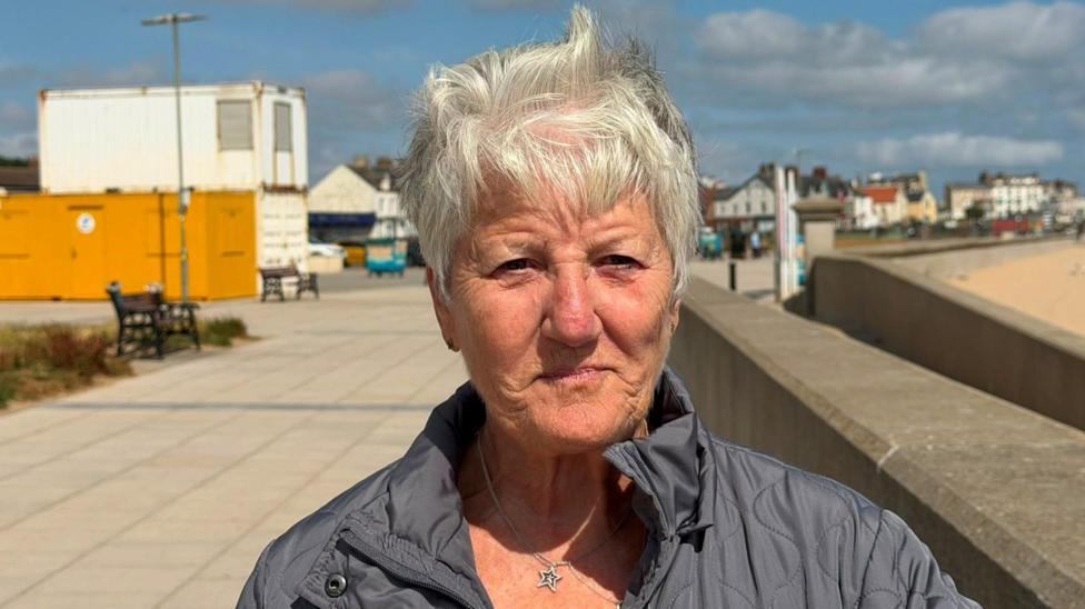Sandra Gleeson has short white hair. She is wearing a black puffer jacket and a silver necklace with a star pendant. 