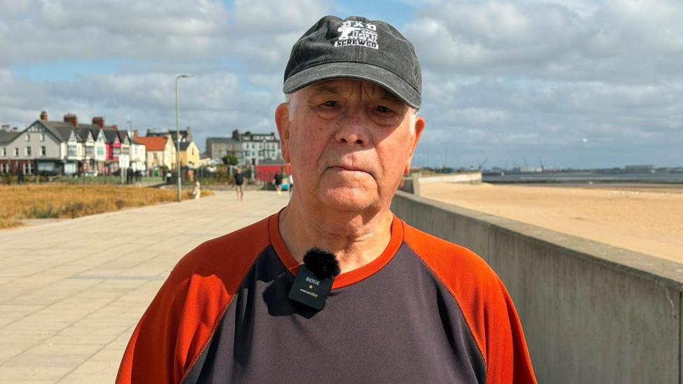 Adrian Barron wears a black cap and a black t-shirt with orange sleeves. He is standing on the promenade next to the sandy beach. 