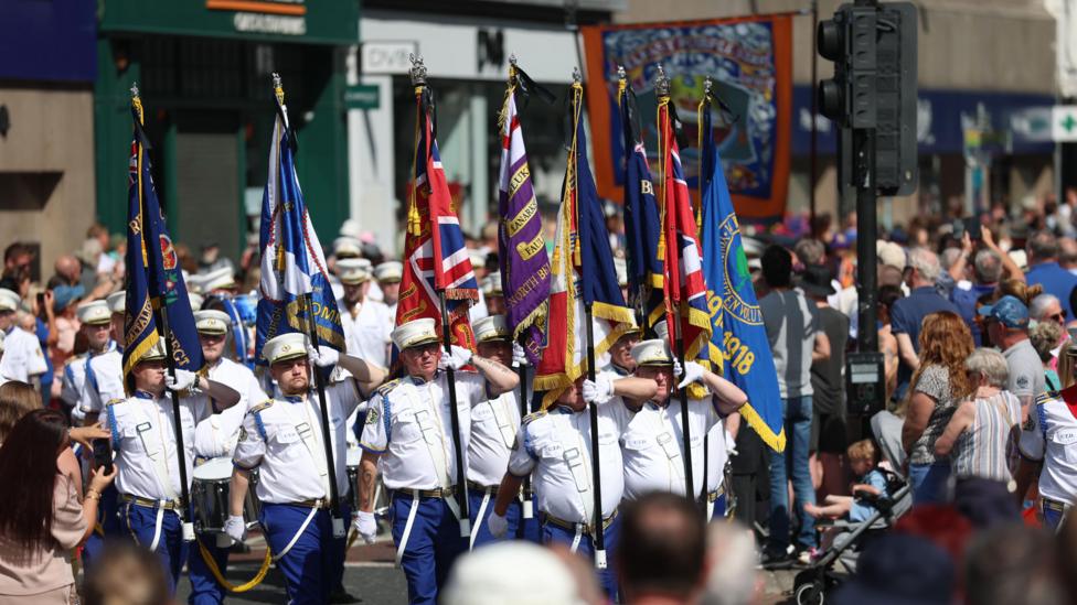 A number of men are walking and holding flags. It is a sunny day and they are wearing white shirts and hats which have a white top and a black peak. There are crowds in the background.
