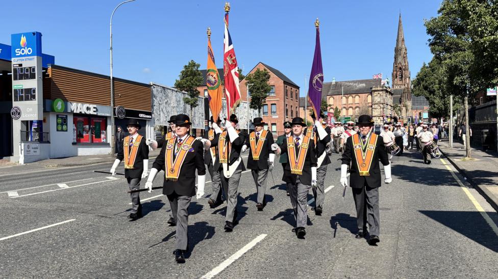 Members of the Orange Order marching. They are each dressed in grey trousers, black coats, white gloves and orange sashes. The have black top hats on. The sky is cloudless and blue.