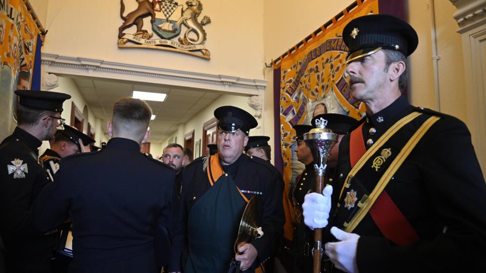 Bandsmen dressed in black with yellow and orange sashes inside Clifton Street Orange hall. On the right of the image one man can be seen holding a brown baton. Several of the men are wearing peaked caps.