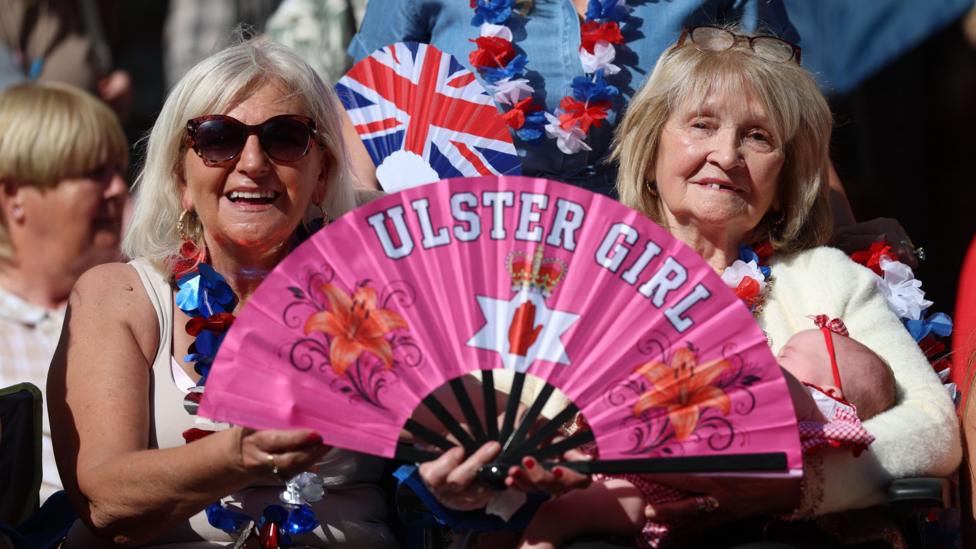 Two women with blond hair are holding up a pink fan that says Ulster girl. One is holding a baby. A crowd is behind them. 