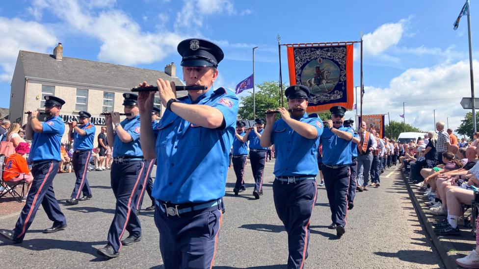 Members of a parade marching in Limavady. Rows of men and women dressed in blue and grey uniform, playing flutes whilst a banner of William III is on display.