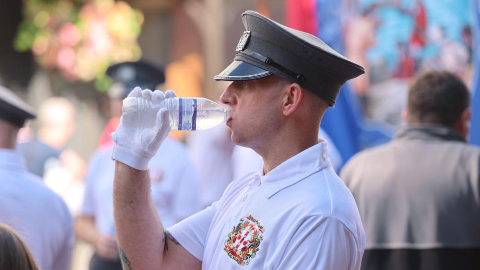 A man in a white shirt drinking from a water bottle. He is wearing a black hat and white gloves and there is a crest featuring a red hand of Ulster symbol on the left breast of his shirt.