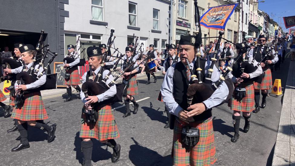 Group of men walking down the high street. They are in orange kilts, blue shirts and black vests. They are playing bagpipes and a banner is flying behind them.