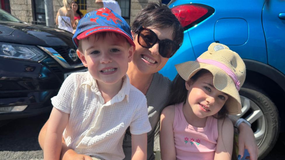 A woman with two children. Woman is wearing sunglasses, and smiling. The little boy has a white shirt and spider-man hat. The girl has a pink sundress and a cream sun hat.