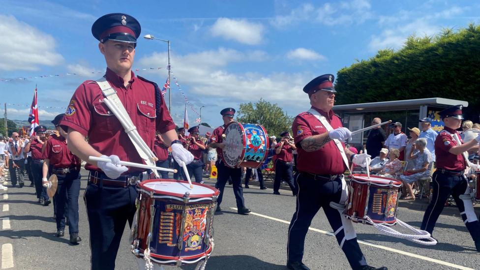 A group of drummers walking on a road. They have a maroon top, navy trousers and a black hat with a black peak. They are playing the drums, with white gloves on and white sticks in their hands.