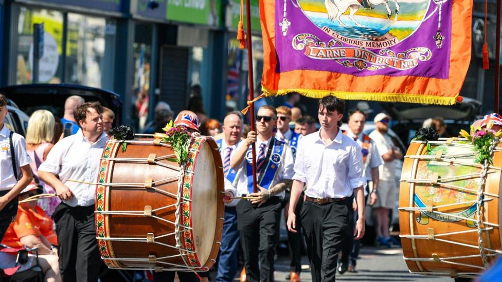 Men holding brown lambeg drums while walking in the parade. Wearing black trousers with light blue shirts, and a blue sash. One man is holding a Larne district orange flag. 