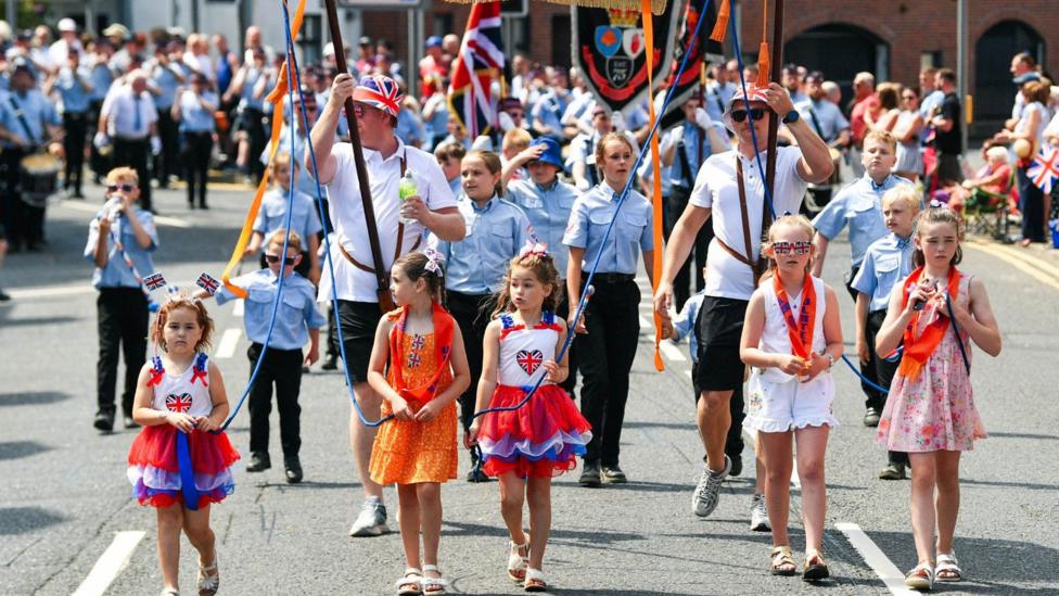 Children and adults walking on a road. Two girls are wearing a red, blue and white dress. Another in an orange dress, and two children in white and pink floral dresses. Behind them there are children in blue shirts and black trouser uniforms.