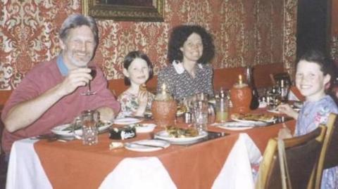 A dinner table with orange table cloth and candles and plates of food. A grey-haired man, brunette woman and two young brunette girls are sat around the table. They are smiling and looking at the camera.
