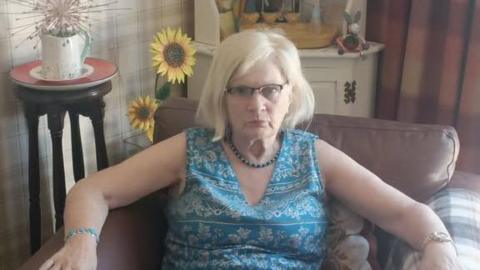 A woman in a blue patterened v-neck dress with bead necklace and turquoise bracelet. She is sitting in a leather chair in a room with checked wallpaper and a tall sideboard behind her with fake sunflowers and ornaments.