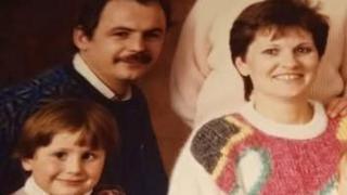 An old family posed photograph featuring a small boy about six, a woman in a cream, pink, yellow, green and brown jumper with short brown hair and a man in a dark jumper with dark hair and moustache.