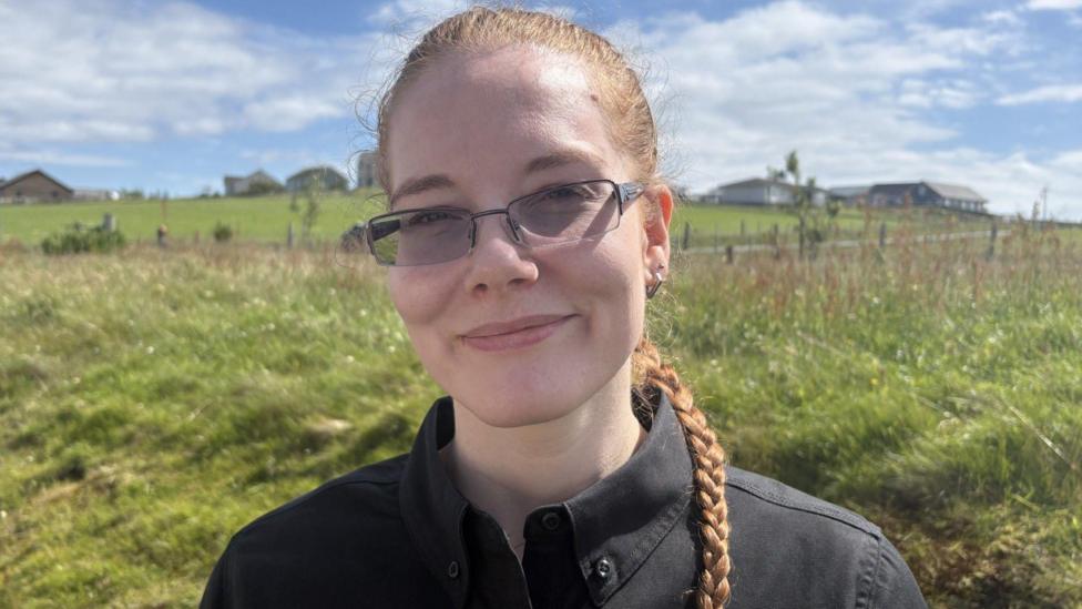 Amy is a young woman in a black shirt. She has long red hair plaited in a single braid and is wearing shaded glasses. She is smiling in a field.