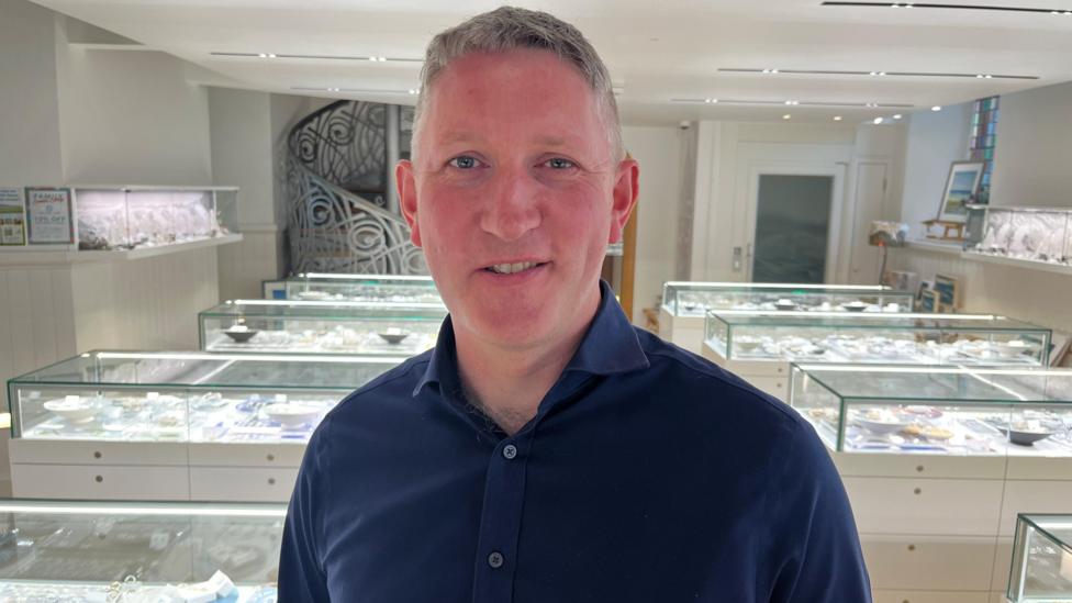 Martin is a grey-haired man in a dark blue open-necked shirt. He is standing in the middle of a jewellery gallery with glass cases behind him.