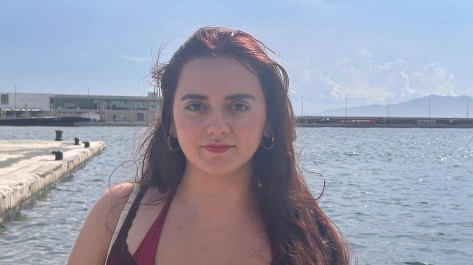 The image is of a young woman with long, dark brown hair falling over her left shoulder. She is looking directly into the camera with a calm expression. She has dark eyebrows, dark eyes, and is wearing a brown top over a burgandy swimsuit, and has small, metallic hoop earrings. Behind her is a wide expanse of calm water with a concrete pier jutting out and several bollards along the edge. In the distance is a long, flat-roofed building and beyond that is a low mountain.