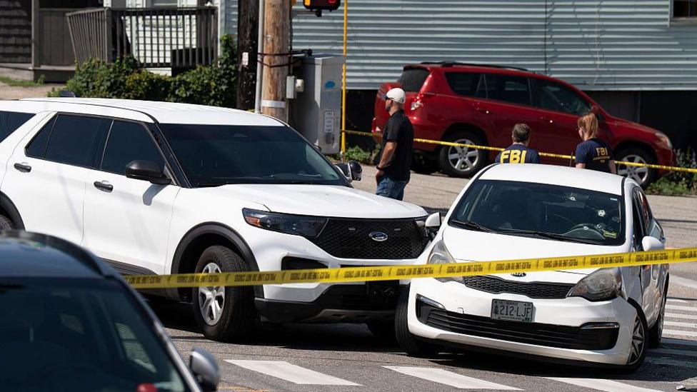 Two white cars that collided are behind yellow police tape. The windscreen of one car has bullet holes in it
