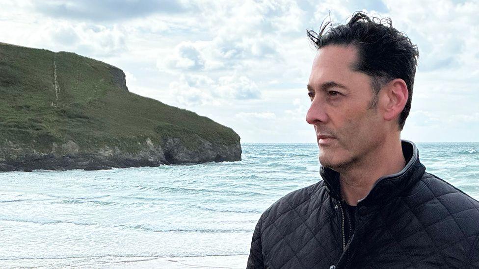 Simon Andriesz wearing a dark quilted jacket stands on a beach beside the sea. The photograph is taken in profile, with Andriesz looking toward the horizon. In the background, waves roll toward the shore and a grassy cliff extends into the water beneath a cloudy sky. The scene is brightly lit by daylight, with the coastline and ocean filling much of the frame.