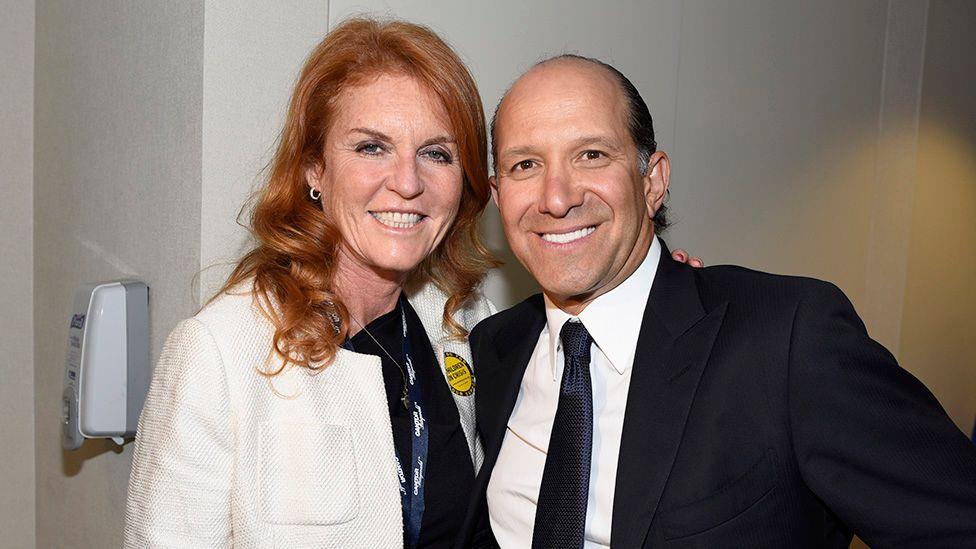 Sarah Ferguson and Howard Lutnick pose together indoors in front of a light-colored wall. One person wears a white textured jacket over a dark top with a lanyard and a round yellow badge, while the other wears a dark suit, white shirt and tie. Both face the camera. A wall-mounted dispenser is visible on the left side of the image.