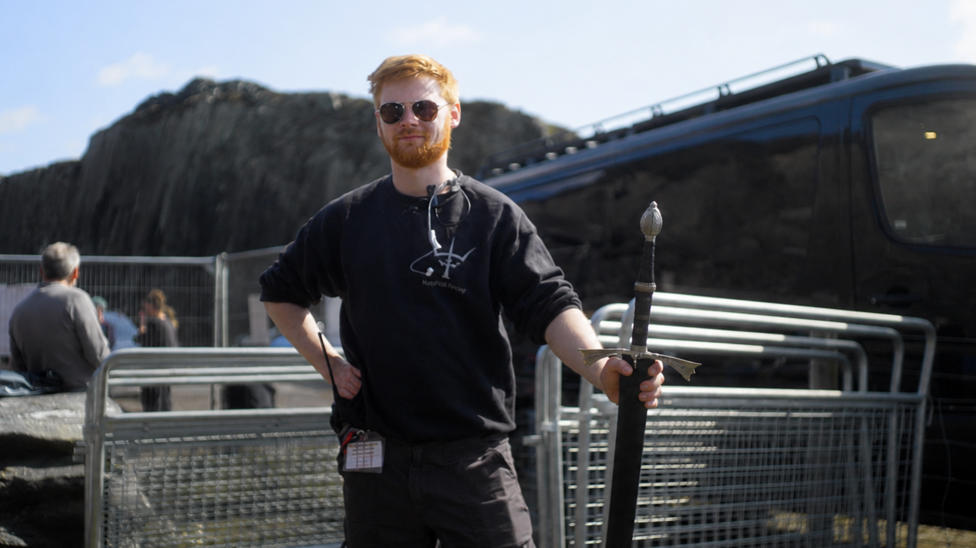 Aaron on set in sunglasses and black top holding a sword, with temporary metal fencing behind him and a truck