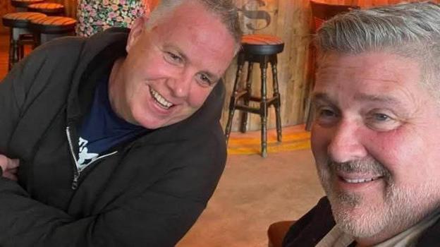 Mike, left, is leaning over towards Terry. He is smiling and wearing a black zip-up hoodie. Terry, right, is looking to the left of the frame and smiling. It is a close up head shot.