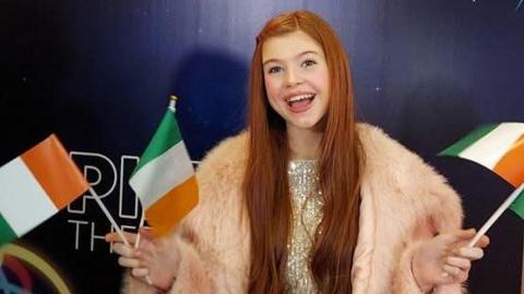 A young girl with long ginger hair. She is holding miniature Irish tricolour flags in both hands. She is wearing a sparkly dress and fur coat.