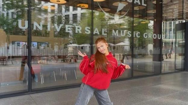 Sophie is standing in front of the Universal Music Group building. She has long ginger hair in a ponytail and is wearing a red hoodie and grey jeans.