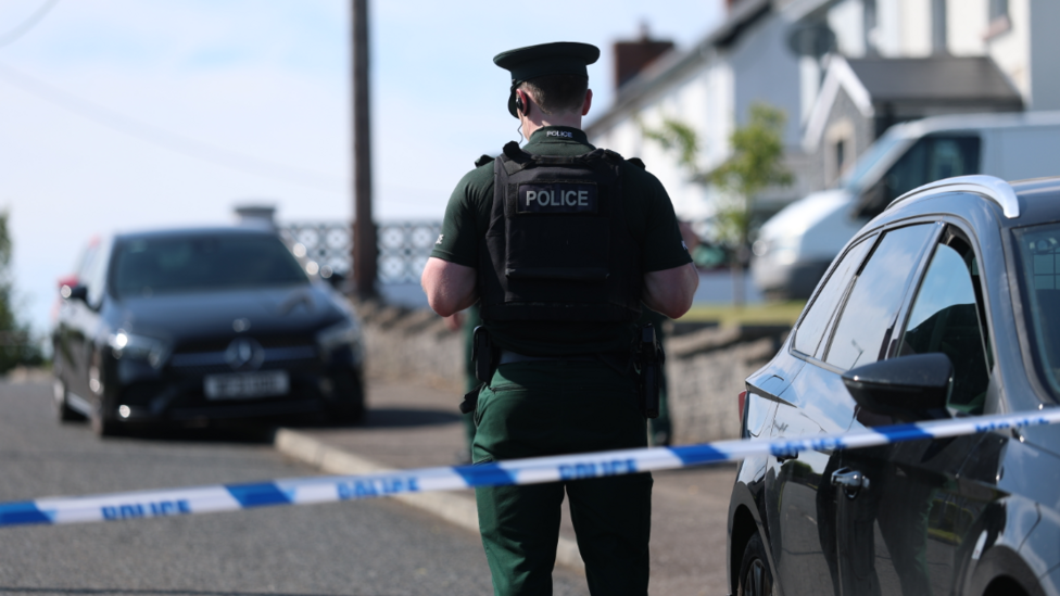 A police officer in full uniform is standing behind a blue and white cordon. This image is in a residential area. There are houses on the right and cars parked outside some o f them.