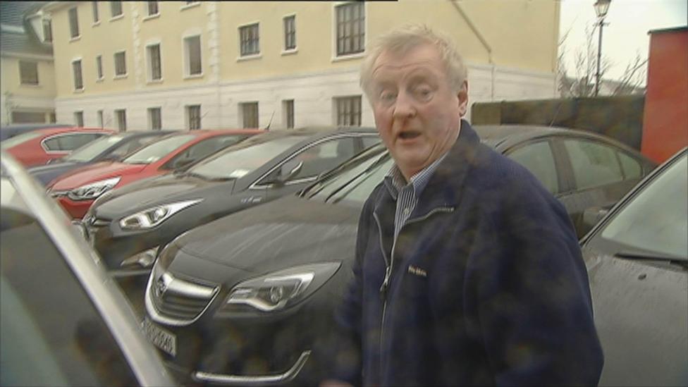 A man with grey hard stands in a car park. He is wearing a blue and white shirt and a navy blue jacket.