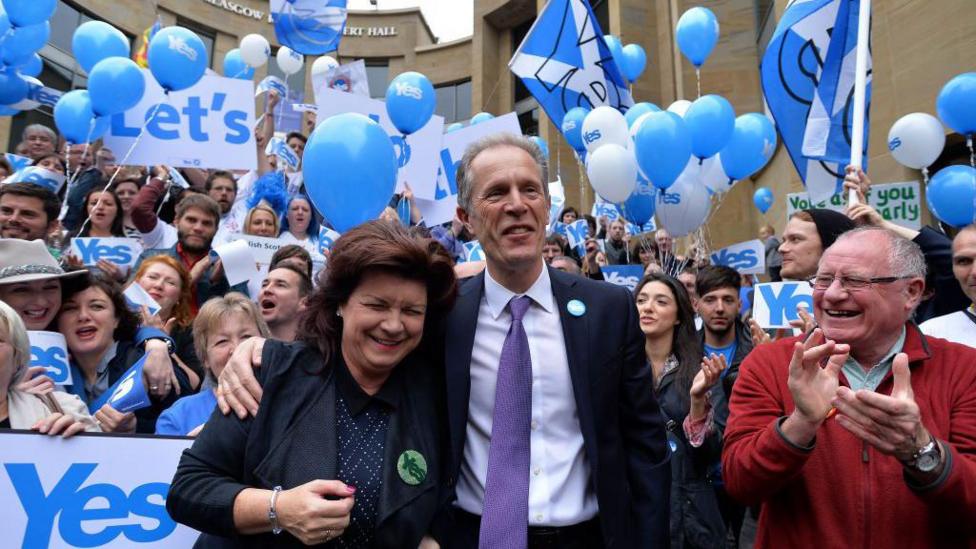 A smiling man in a suit, shirt and tie has his arm round the should over a smiling woman as they stand in front of cheering crowds