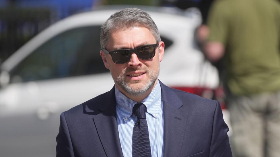 Adam Smyth, Director for BBC Northern Ireland, wears black Ray Ban sunglasses. he has short greying hair and a beard. He wears a navy suit, blue shirt and navy tie.