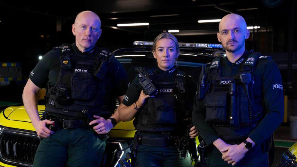 Three police officers looking at the camera. All are wearing police uniform that is black and dark green. The one on the left is a bald man, in the middle is a blonde woman and on the right is a bald man with dark beard.