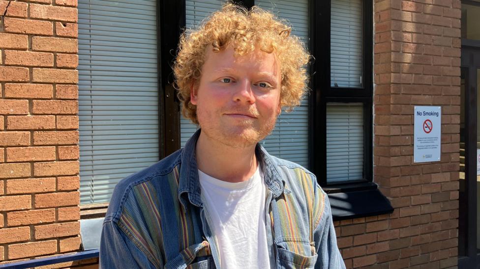 A man with curly ginger hair and ginger stubble standing next to a blue railing.