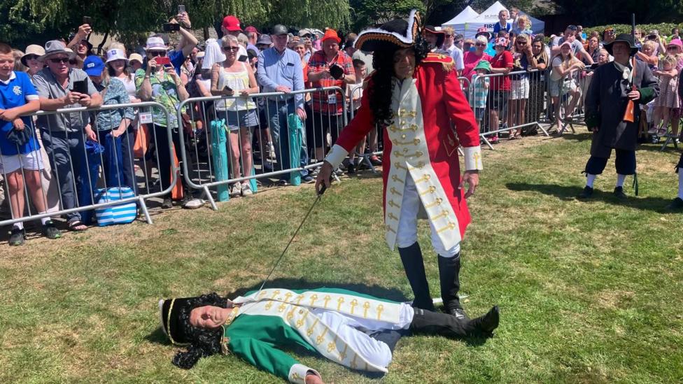 A man dressed like King William III is standing over a man dressed like King James II who is lying on grass with his eyes closed. William has his sword pointed towards James. A crowd is watching. 