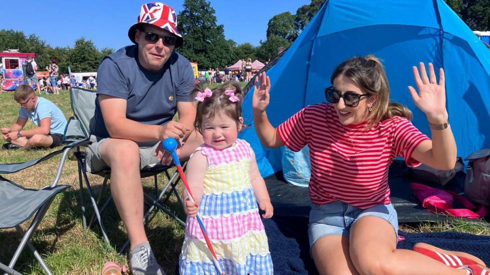 A toddler is smiling. She's holding a blue, red and white baton. She's wearing a pink, white, yellow and blue dress. A crowd is in the background. 