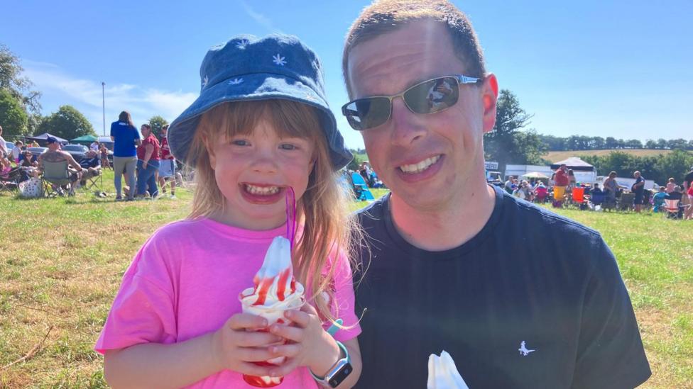 A girl is smiling. She is holding an ice cream in a plastic cup. Strawberry sauce is on top. A man next to her has an ice cream in a cone. He is wearing sunglasses. 