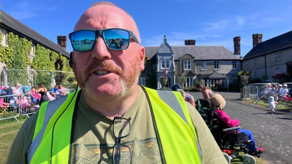 A man is wearing a bright yellow high viz jacket. He has sunglasses on and glasses are folded around the nape of his top. A design featuring a mountain range is on his olive coloured top. People are behind him in the background. A two-story country house is behind him. 