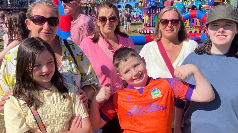 Three women wearing sunglasses and three children are standing together. The boy in the middle is holding his arms up. A fun fair is behind them. 