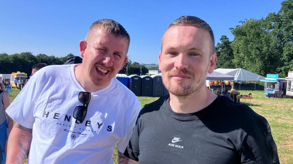 Two men are standing together. One has a white top with sunglasses folded over the nape of his top. The other is wearing a black top. Portaloos are in the background. 