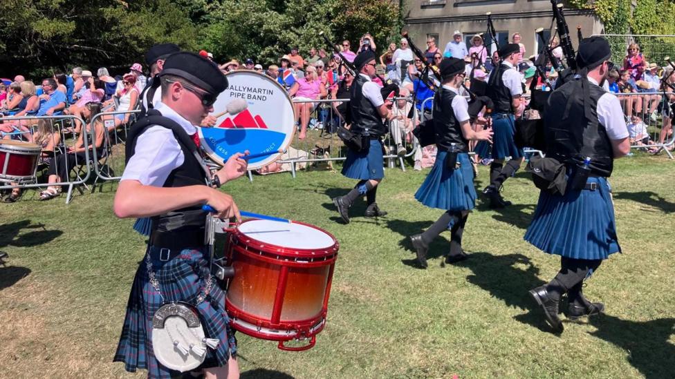 A parade with men playing instruments. A boy in the foreground is playing the drums. Men marching in front of him are playing pipes. A crowd is watching. 