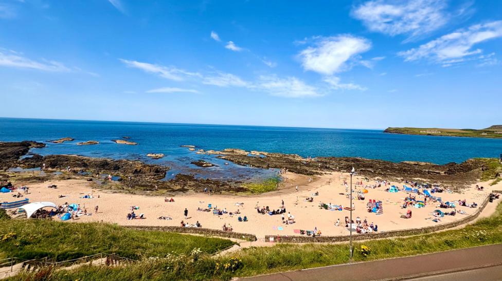 A wide landscape picture of a beach crowed with people. The sea and the sky are blue and there are only a few whispy clouds.