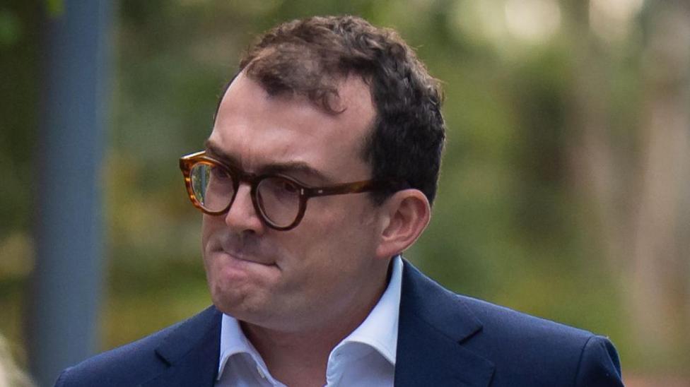 A man who has short dark hair, glasses and who is wearing a navy suit jacket and white shirt. He is looking away from the camera.
