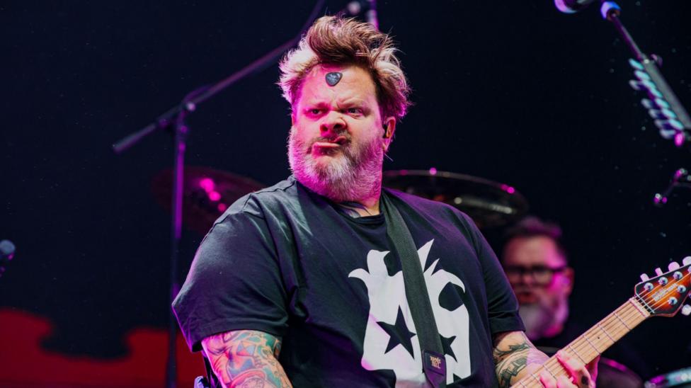 Jaret Reddick is pictured whilst performing on stage at Cardiff Castle. He is wearing a black t-shirt with a white graphic print in the centre of it. He has brown hair, tattoos on his arms, and is holding a guitar. On the middle of his forehead is a guitar pick.