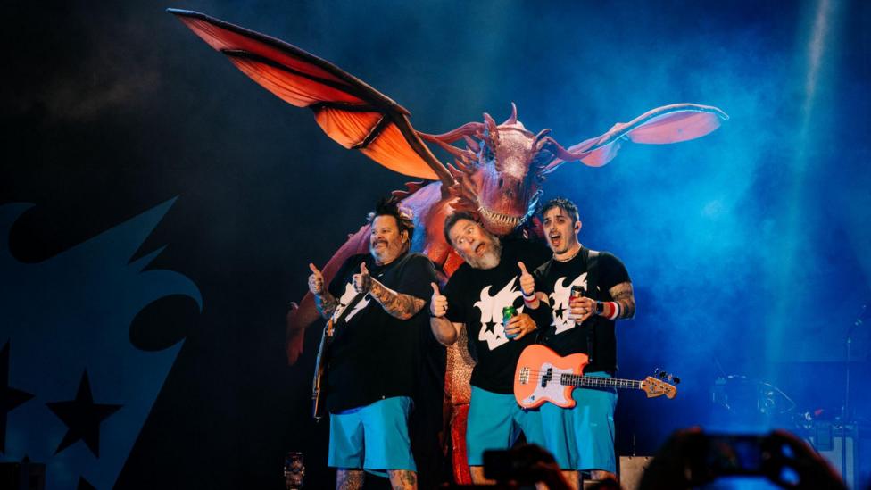 Frontman Jaret Reddick, bassist Rob Felicetti and drummer Gary Wiseman, from the band Bowling for Soup. The three band members are stood on stage whilst performing at Cardiff Castle. Behind them is a large red dragon, appearing over their heads. The band are all holding their thumbs up. Reddick and Felicetti are wearing guitars. All three members are wearing blue shorts and a black t-shirt with a white graphic print in the centre.
