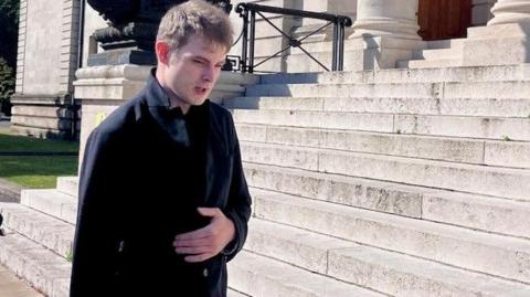 A man with light short hair and a dark coat walks up light stone steps, with his head down.
