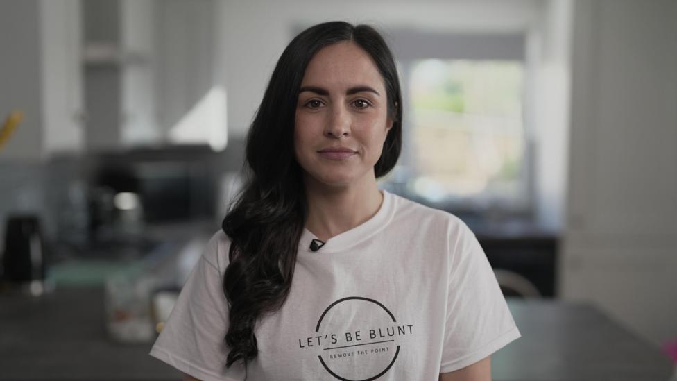 Leanne Lucas, who has long wavy dark hair, and is wearing a white t-shirt with a circular logo with the catchphrase 'Let's Be Blunt' on the front. She is standing in a kitchen looking into the camera with a determined expression.