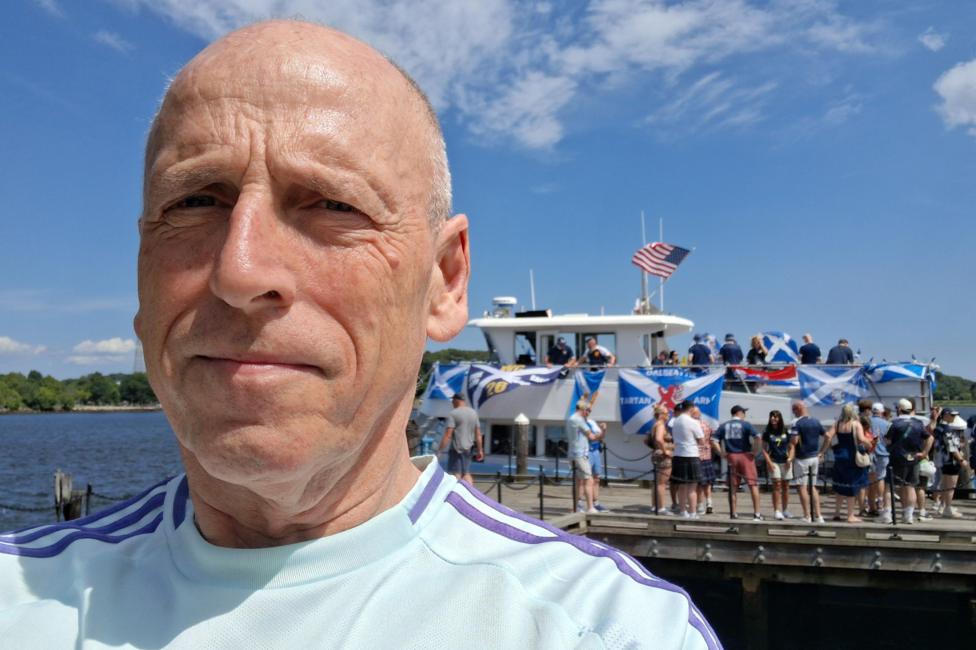 Hamish Husband, wearing a light blue Scotland top, takes a selfie in front of a group of Scotland fans boarding a saltire-clad boat. There is a clear blue sky and water in the background