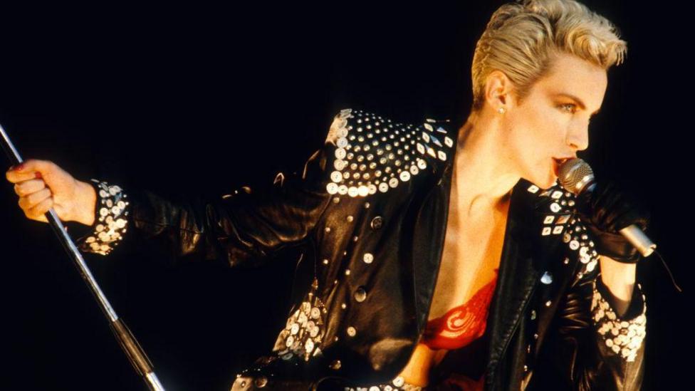 Annie Lennox singing into a mic during the 1980s. She has short, cropped, blonde hair and is wearing a black leather jacket with sequins attached