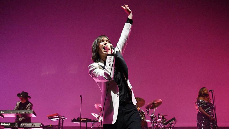 Primal Scream performing on stage. Singer Bobby Giillespie has shoulder-length, dark hair and is wearing a light suit jacket over dark clothes. He is singing into a mic with his arm raised in the air. 