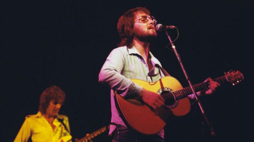 Gerry Rafferty has a beard and is wearing glasses as he plays guitar and sings on stage 